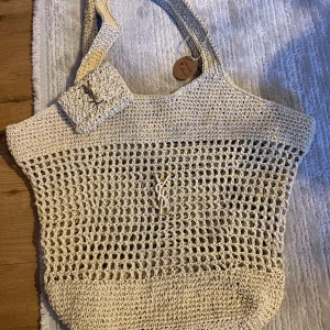 Beige stickad handväska från Yves Saint Laurent - Säljer en beige stickad handväska från Yves Saint Laurent med matchande liten väska. Väskan har ett öppet nätmönster och YSL-logga i metall framtill. Perfekt för att bära med sig det viktigaste på stan eller stranden.