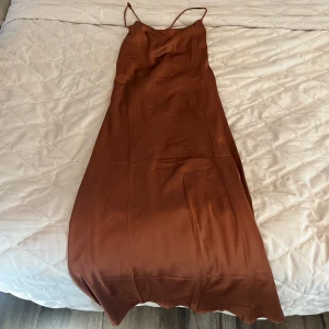 Brun långklänning med smala axelband - Säljer en snygg brun långklänning med smala axelband och omlottknytning i ryggen. Klänningen har en elegant, slät yta och faller fint längs kroppen. Perfekt för dig som gillar minimalistisk och stilren design.