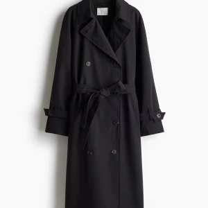 Svart trenchcoat med bälte - Snygg svart trenchcoat med klassisk dubbelknäppning och bälte i midjan. Jackan har långa ärmar med knappar vid ärmslut och bred krage. Perfekt för en stilren look under kyligare dagar.