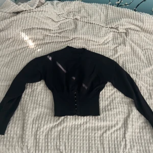 Svart croppad kofta med knappar - Säljer en svart croppad kofta med långa ärmar och knappar framtill. Tröjan har en tight passform vid midjan och är tillverkad i ett stickat material. Perfekt att styla till olika outfits!