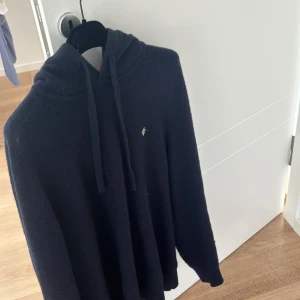 Mörkblå stickad hoodie från Morris - Säljer en mörkblå stickad hoodie från Morris i storlek M. Tröjan har huva med dragsko och en liten broderad logga på bröstet. Perfekt för dig som gillar klassisk och stilren design.