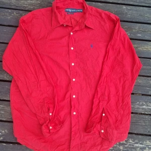 Röd skjorta från Ralph Lauren - Säljer en klassisk röd skjorta från Ralph Lauren i bomull. Skjortan har lång ärm, knappar framtill och en blå broderad logga på bröstet. Modellen är Classic Fit och passar perfekt till dig som gillar en tidlös stil.