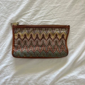 Mönstrad kuvertväska med bruna detaljer - Säljer en snygg kuvertväska från Missoni med ikoniskt mönster i jordnära färger som brunt, grönt, gult och orange. Väskan har dragkedja upptill och bruna kanter i skinn. Perfekt accessoar för att lyfta din outfit!