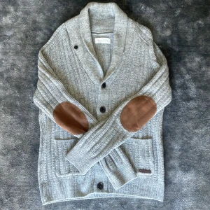 Grå stickad kofta från Pier One - Säljer en grå stickad kofta från Pier One med bruna mockadetaljer på armbågarna. Koftan har även fickor framtill och är i nyskick! Storlek M