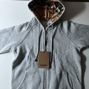 Grå hoodie från Burberry med dragkedja - Säljer en grå hoodie från Burberry med klassiskt rutigt foder i huvan och dragkedja framtill. Tröjan har fickor och justerbar huva med snören. storlek s/m. Pris kan inte sänkas.