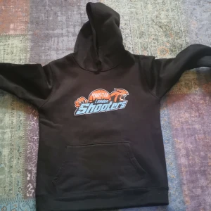 Svart hoodie trapstar London shooters  - Svart hoodie med stor ficka framtill och tryck av London Shooters i orange, blått och vitt på bröstet. Klassisk huva och mjukt material, perfekt för en sportig look.