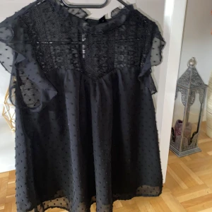 Svart blus med spets från SHEIN - Säljer en svart blus från SHEIN med transparenta, korta volangärmar och spetsdetaljer upptill. Blusen har ett diskret prickigt mönster i tyget och en lätt, luftig känsla. Perfekt för dig som gillar romantiska detaljer och svart bas.