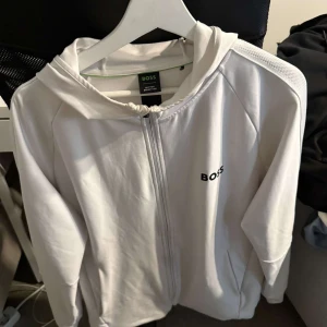 Vit zip-hoodie från BOSS - Säljer en stilren vit hoodie med dragkedja från BOSS. Jackan har huva, långa ärmar och en diskret BOSS-logga på bröstet. Detalj med rand på ena ärmen. Perfekt för en clean och sportig look.
