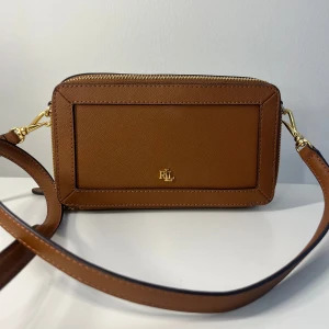 Ralph Lauren axelremsväska  - Superfin äkta axelremsväska från Ralph Lauren. Den har aldrig blivit använd och är i superfint skick💕Väskan är gyllenbrun, med gulddetaljer (på loggor, kedjor och spännen). Andra bilden visar en närbild på loggan och bild 3 visar den tryckta Ralph Lauren loggan inne i väskan. RLL finns ingraverat på spännet. Väskan har två lite större fack. Det ena har även lite mer fack inuti. Det andra facket är indelat i två. YKK finns ingraverat på dragkedjan. Skriv gärna för fler bilder eller vid frågor☺️
