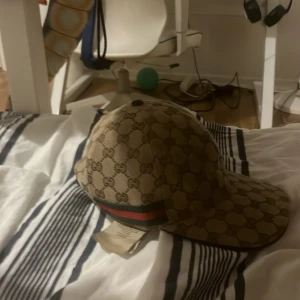 Gucci keps med monogram och röd-grön rand - Säljer en beige keps från Gucci med klassiskt GG-monogram över hela kepsen och en röd-grön rand på sidan. Justerbar rem baktill. Perfekt för dig som gillar lyxiga accessoarer och vill sticka ut.