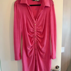 Rosa ribbad skjortklänning med knappar - Säljer en rosa ribbad skjortklänning med långa ärmar och krage. Klänningen har knappar hela vägen framtill och snygg rynkdetalj i mitten som ger en cool effekt. Perfekt för dig som vill sticka ut lite extra!