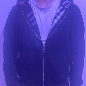 Svart hoodie med dragkedja och mönstrad huva från Burberry - Säljer en svart hoodie med dragkedja från Burberry. Har använts ganska länge typ 1 år säljer för den är för lite på mig. Model är 175 cm och 70 kg. Pris kan diskuteras vid snabb affär 