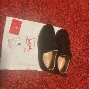 Svarta sneakers med nitar från Christian Louboutin - Säljer ett par svarta sneakers från Christian Louboutin med ikoniska röda sulor och svarta nitar på tån. Skorna har snörning och är tillverkade i skinn. Kommer med originalpåse och extra skosnören.