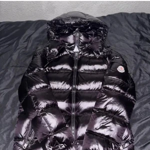 Svart dunjacka från Moncler med huva - Säljer en glansig svart dunjacka från Moncler med huva och dragkedja framtill. Jackan har lång ärm och klassisk Moncler-logga på ärmen. Perfekt för kyliga dagar och riktigt snygg streetstil.