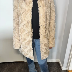 Beige pälsjacka med knappar - Säljer en beige pälsjacka med snyggt mönstrad struktur och klassisk krage. Jackan har långa ärmar och stängs med knappar framtill. Perfekt för dig som vill ha en mysig och trendig look.