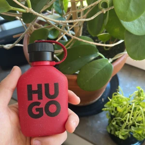 HUGO röd parfymflaska - Säljer en parfym från HUGO i en matt röd flaska med svart skruvkork och röd snodd. Flaskan har en fyrkantig form och stor svart text på framsidan. Perfekt för dig som gillar stilrena och moderna dofter. Flaskan är helt full!