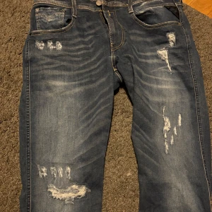 Blå slitna jeans med raka ben - Säljer ett par blå jeans med slitningar och trasiga detaljer på låren och knäna. Jeansen har normal passform och raka ben, klassisk femficksmodell och knappgylf. Perfekta för en avslappnad stil. Storlek 32 och i perfekt skick helt nya!