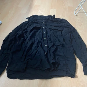 Svart skjorta från Gina Tricot - Säljer en svart skjorta från Gina Tricot med klassisk krage och knäppning framtill. Skjortan har långa ärmar och är tillverkad i ett lätt och mjukt tyg, perfekt för en stilren look.