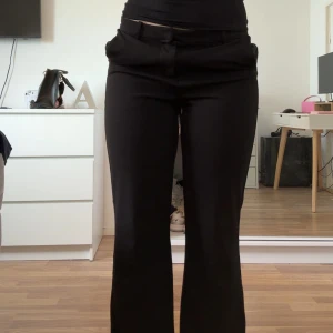 Svarta kostymbyxor med bootcut - Svarta kostymbyxor med bootcut-snitt och låg midja. Byxorna har klassisk passform och bälteshällor i midjan. Perfekta för en stilren look och passar till många olika outfits.