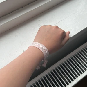 Rosa armband från Dior - Säljer ett ljusrosa armband från Dior med vit broderad text och tofsar i ändarna. Armbandet är justerbart och har en snygg, ungdomlig stil som passar perfekt som accessoar.