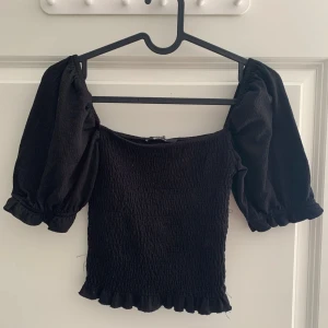 Svart croppad topp med puffärmar - Trendig svart croppad topp med puffiga kortärmar och smockad, stretchig passform. Fyrkantig ringning och volangkant. Storlek S. Köpt på Gina Tricot. Mycket använd med en tråd som lossnat. Inget som påverkar funktionen.