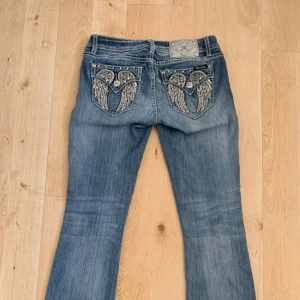 Miss Me jeans W27 med broderade fickor och bootcut - Snygga blå jeans från Miss Me med bootcut-modell och broderade bakfickor med vingar och nitar. Jeansen har slitningar framtill och dekorativa detaljer vid fickorna.o Perfekta för dig som vill sticka ut med coola detaljer. Köpta på vinted men alldeles för små för mig. 38 cm tvärs över i midjan. 75cm innerbenslängd. 