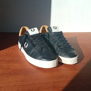 Svarta sneakers från Fred Perry - Säljer ett par svarta Fred Perry sneakers med vit sula och beige insida. Skorna har snörning och klassisk Fred Perry-logga på sidan och plösen. Perfekta för dig som gillar stilrena och sportiga skor. Lite creasade där fram.