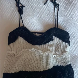 Svart och vit stickad topp med spetsdetaljer - Säljer en unik stickad tröja i svart och vitt med smala axelband som knyts. Tröjan har dekorativa spetsdetaljer och en vågig kant upptill. Perfekt för dig som gillar en bohemisk och trendig stil.