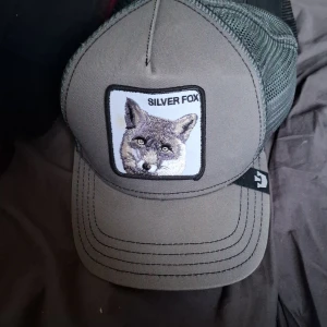 Grå keps med broderad räv - Snygg grå keps från Goorin Bros med broderad patch framtill som föreställer en räv och texten 'SILVER FOX'. Kepsen har nät baktill och böjd skärm för en klassisk look.