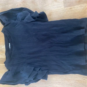 Svart ribbad topp med volangärmar - Säljer en svart topp med ribbad struktur och volangdetaljer på axlarna. Toppen har rund halsringning och korta ärmar med extra volym. Perfekt för dig som vill ha något stilrent men ändå med en twist. I storlek 146/152 men passar mgo med storlek 158/164. Köparen står för frakt!