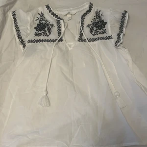 Vit blus med broderade detaljer - Säljer en vit blus med svarta broderade blommor och dekorativa band framtill. Blusen har korta ärmar med broderad kant och knytband med tofsar vid halsen. Perfekt för en somrig och bohemisk stil.