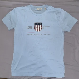 Ljusblå t-shirt från GANT - Säljer en ljusblå t-shirt från GANT med tryckt logga och sköld på bröstet. T-shirten har rund halsringning och korta ärmar. Perfekt för en avslappnad och stilren look.