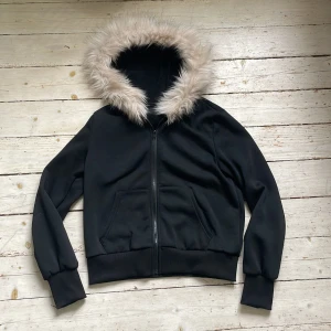 Svart bomberjacka med fuskpäls - Svart bomberjacka med dragkedja och huva kantad med ljus fuskpäls. Jackan har långa ärmar, ribbade muddar och två fickor framtill. Perfekt för kyligare dagar.