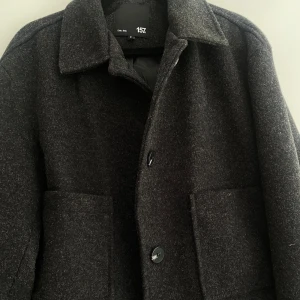 Mörkgrå ullkappa från 157 - Stilren mörkgrå kappa i ull från 157 med klassisk krage och stora framfickor. Kappan har knäppning framtill med svarta knappar och en enkel, rak passform. Perfekt för kyliga dagar och ger en tidlös look. Kappan är använd 1 gång bara så den är i helt nytt skick! 