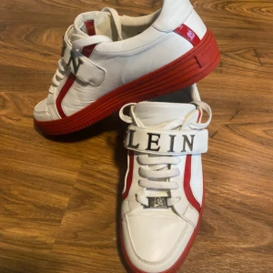 Vita och röda sneakers från Philipp Plein - Säljer ett par vita sneakers från Philipp Plein med röda detaljer och röd sula. Skorna har snörning, bred kardborre med texten 'PLEIN' och metallplatta vid snörningen. Tillverkade i skinn med en sportig och modern look.
