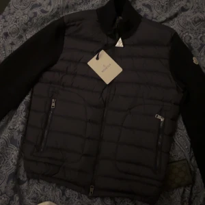 Svart dunväst från Moncler med ärmar - Säljer en svart dunväst från Moncler med långa svarta ärmar och ståkrage. Västen har två framfickor med dragkedja och Moncler-logga på ärmen. Perfekt för kyliga dagar. Storlek M men passar S också 