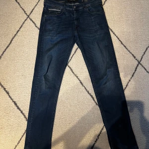 Replay Hyperflex jeans i mörkblå denim - Säljer ett par Replay Hyperflex jeans i mörkblå tvätt med klassisk femficksdesign och raka ben. Jeansen har snygga kontrastsömmar och en diskret Replay-logga på bakfickan. Perfekta för dig som gillar stilrena och bekväma jeans. Storlek L34 W34, modellen grover.