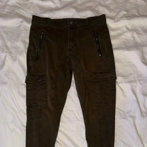 Bruna cargopants från Jack's Studio Collection - Säljer ett par bruna cargopants från Jack's Studio Collection i storlek 46. Byxorna har flera fickor med lock på sidorna, dragkedjor framtill och förstärkta sömmar vid knäna. Perfekta för dig som gillar en avslappnad och funktionell stil.