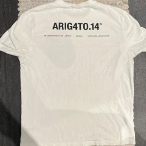 Axel Arigato T-Shirt Vit - Vit t-shirt från Axel Arigato, storlek M, i mycket fint skick. Stilren med ett litet tryck på bröstet och ett större på ryggen – modern och trendig design i hög kvalitet.