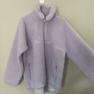 Ljuslila fleecejacka med dragkedja - Mysig ljuslila fleecejacka med hög krage och hel dragkedja framtill. Jackan har två fickor med dragkedja och är långärmad. Perfekt för kyliga dagar när du vill vara både varm och stilren.