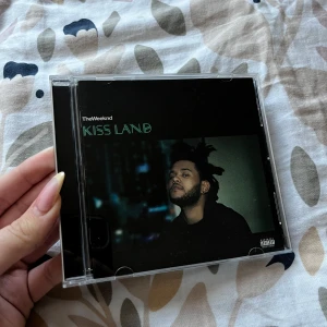 Kiss Land - Säljer CD-albumet 'Kiss Land' av The Weeknd. Omslaget är svart med ett porträtt av artisten på framsidan och en grön grafisk design på baksidan och skivan. Perfekt för dig som samlar på musik eller gillar The Weeknd.