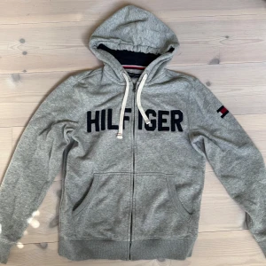 Grå hoodie från Tommy Hilfiger - Grå hoodie med dragkedja från Tommy Hilfiger. Av hög kvalité! Säljer pga för infrekvent användning.