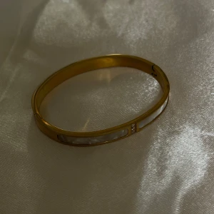 Guldarmband med vita detaljer - Elegant guldfärgatarmband med vita detaljer och en stilren design. Armbandet har en slät yta med inlägg av vit pärlemor och små glittrande stenar. Perfekt för att ge en touch av lyx till din outfit.