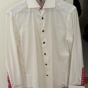 Vit skjorta från La Chemise - Säljer en stilren vit skjorta från La Chemise med rödrutiga detaljer på insidan av kragen och ärmsluten. Skjortan har svarta knappar och är långärmad. Perfekt för en elegant look med en twist. 🕴️