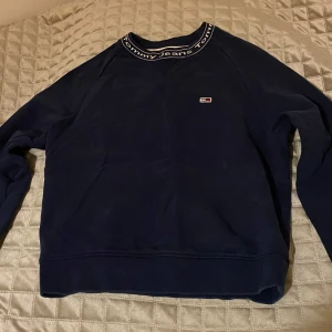 Mörkblå tröja från Tommy Jeans - Säljer en snygg mörkblå tröja från Tommy Jeans med deras ikoniska logga på bröstet. Tröjan har en rund halsringning med textdetaljer och långa ärmar. Perfekt för en stilren och avslappnad look.