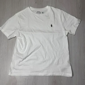 Vit t-shirt från Ralph Lauren - Säljer en klassisk vit t-shirt från Ralph Lauren med det ikoniska broderade logotypen på bröstet. T-shirten är kortärmad och tillverkad i mjuk bomull för en bekväm passform. Perfekt för en stilren och avslappnad look.