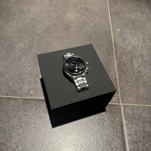 Silverfärgad klocka från BOSS - Snygg silverfärgad klocka från BOSS med svart urtavla och kronografdetaljer. Armbandet är i rostfritt stål och klockan levereras i en elegant svart förpackning med BOSS-logga. Perfekt för den stilmedvetna! Nypris 5000kr