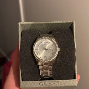 Silverfärgad klocka från Guess - Snygg silverfärgad klocka från Guess med romerska siffror. Urtavlan är omgiven av glittrande stenar och armbandet är i rostfritt stål. Perfekt för att ge en elegant touch till din stil. (Box tillgår)