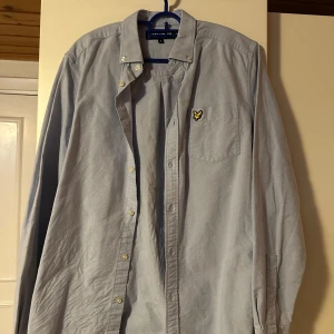 Ljusblå skjorta från Lyle & Scott - Säljer en stilren ljusblå skjorta från Lyle & Scott med klassisk krage och knappstängning. Skjortan har en broderad logotyp på bröstfickan och långa ärmar. Perfekt för en avslappnad men ändå uppklädd look.