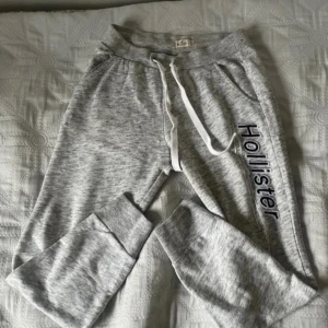 Grå mjukisbyxor från Hollister - Säljer ett par grå mjukisbyxor från Hollister med snörning i midjan och ribbade muddar vid bensluten. Byxorna har en avslappnad passform och Hollister-loggan längs benet. Perfekta för en bekväm dag hemma eller en avslappnad utflykt.  Har ett pyte litet fläck som syns knappt går någ säkert bort i tvätten
